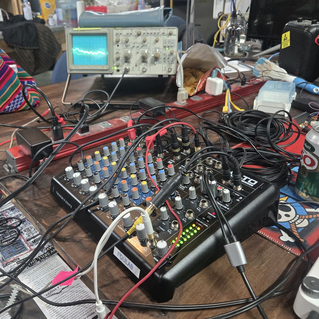 A wooden table with a mixer, wires, and an oscilloscope in the background displaying a wave representing the sound coming out of the mixer.