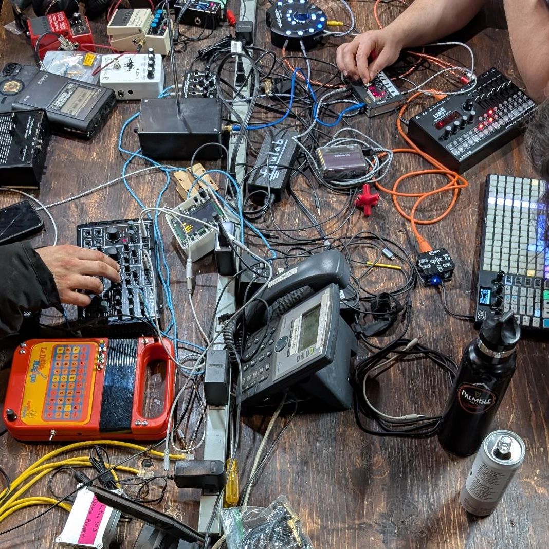 A color photo of musical equipment, wires, and hands on top of a wooden table