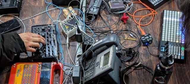 A color photo of musical equipment, wires, and hands on top of a wooden table
