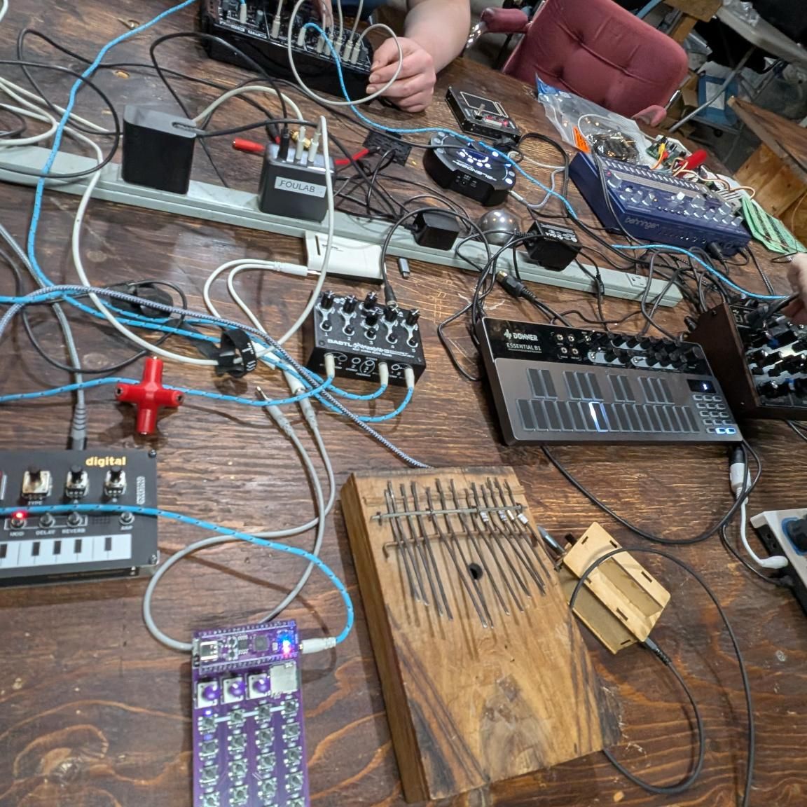 A wooden table filled with electronic music equipment and a kalimba. An outstretched arm adjusting a setting on a synth.